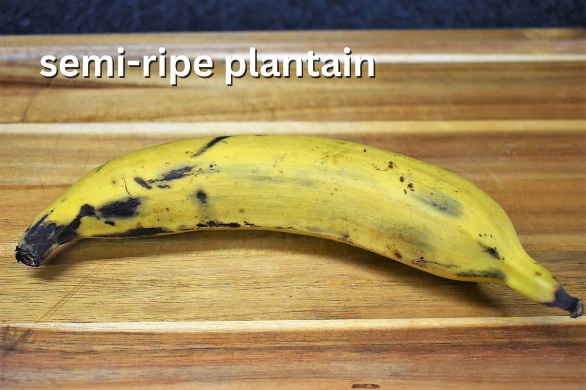 A whole yellow plantain with some dark spots on a wooden cutting board, labeled "semi-ripe plantain."